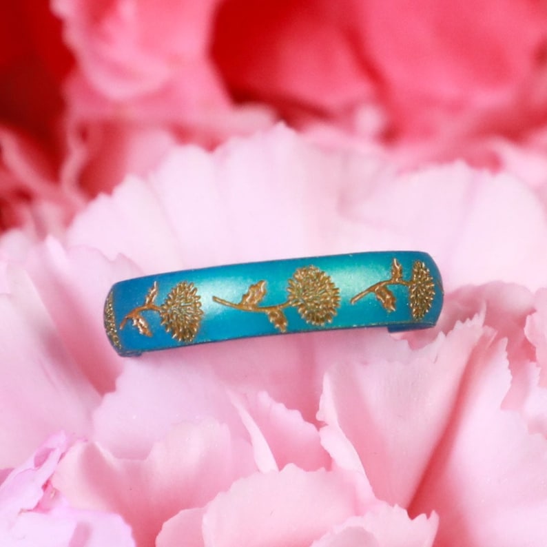 May include: A turquoise blue ring with a gold floral design. The ring is set against a pink background.