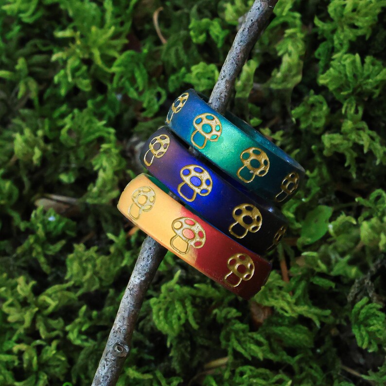 May include: Four colorful rings with mushroom designs. The rings are in shades of blue, green, purple, yellow, and red. Each ring has a gold-colored mushroom pattern. The rings are stacked on a small branch against a background of green moss.