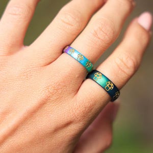 May include: Two rings with a mushroom design. One ring transitions from purple to blue, the other from green to blue. Both rings have a gold mushroom pattern. The rings are on a person's finger.