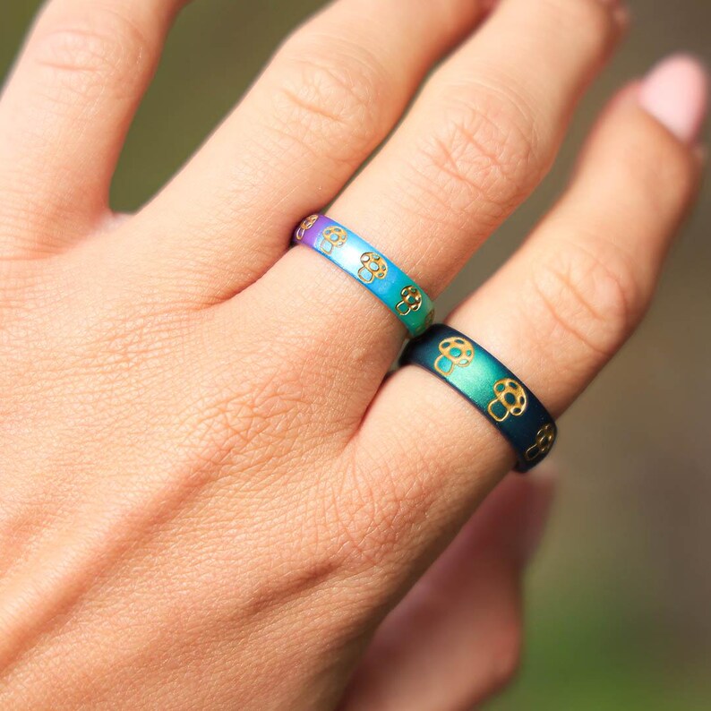 May include: Two rings with a mushroom design. One ring transitions from purple to blue, the other from green to blue. Both rings have a gold mushroom pattern. The rings are on a person's finger.