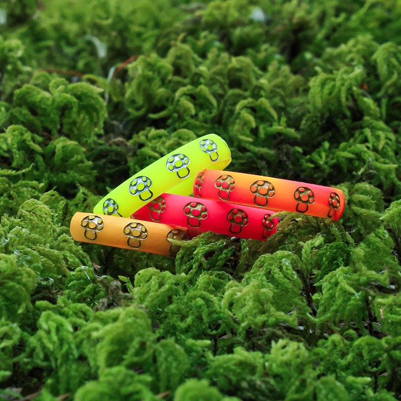 May include: A collection of colorful, cylindrical objects with a mushroom design. The objects are in shades of yellow, orange, and red, and are set against a backdrop of green moss. The mushroom design is in black.