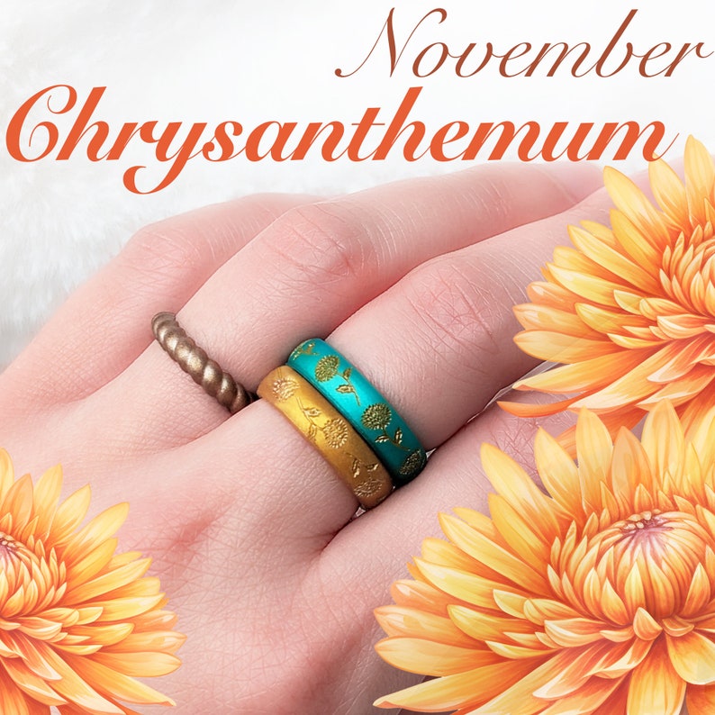 May include: Two silicone rings with floral designs. The top ring is turquoise with gold floral accents. The bottom ring is gold with a darker gold floral design. The rings are worn on a hand with a background of orange chrysanthemums.