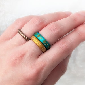 May include: Two silicone rings stacked on a finger. The top ring is turquoise blue with a gold floral pattern. The bottom ring is gold with a gold floral pattern.