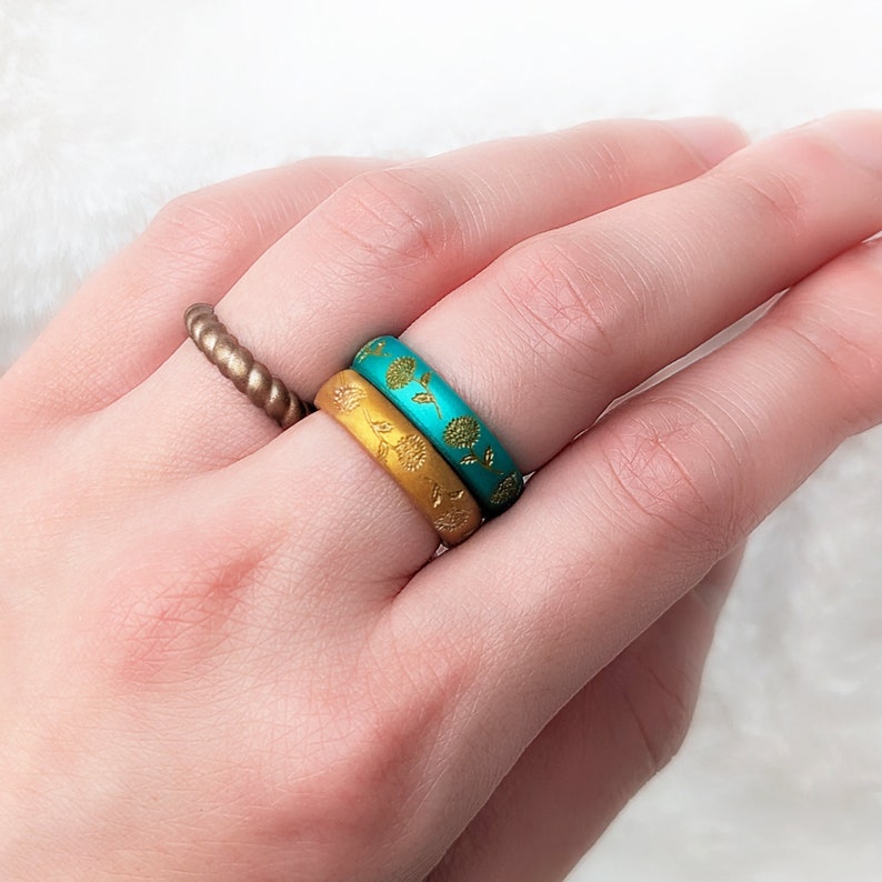 May include: Two silicone rings stacked on a finger. The top ring is turquoise blue with a gold floral pattern. The bottom ring is gold with a gold floral pattern.