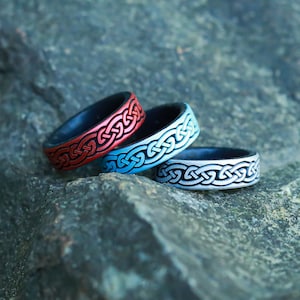 May include: Three silicone rings with a Celtic knot design. The rings are stacked on top of each other. The top ring is silver, the middle ring is blue, and the bottom ring is red.