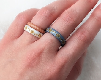 Könnte beinhalten: Zwei Silikonringe mit Blumenmuster. Der obere Ring ist weiß mit goldenen Blumen und der untere Ring ist blau mit goldenen Blumen.