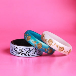 May include: Three decorative bangles stacked on top of each other. The top bangle is white with gold floral designs. The middle bangle is teal blue with gold floral designs. The bottom bangle is light purple with black floral designs.