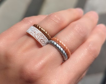Könnte beinhalten: Drei Silikonringe, die auf einem Finger übereinander liegen. Der obere Ring ist weiß mit einem Blumenmuster, der mittlere Ring ist kupferfarben und der untere Ring ist silberfarben mit einer pfirsichfarbenen Basis.