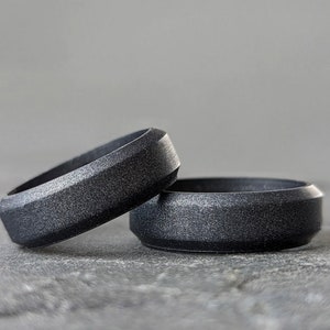 Op de afbeelding: Twee donkergrijze ringen met een getextureerd oppervlak en een geometrisch ontwerp. De ringen zijn gemaakt van een mat materiaal en worden gepresenteerd op een grijze ondergrond. De ringen zijn een moderne interpretatie van een klassiek accessoire.