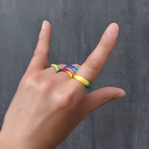 Puede incluir: Una mano con cuatro anillos de silicona de colores. Los anillos est&aacute;n apilados uno encima del otro y son de los colores del arco&iacute;ris.