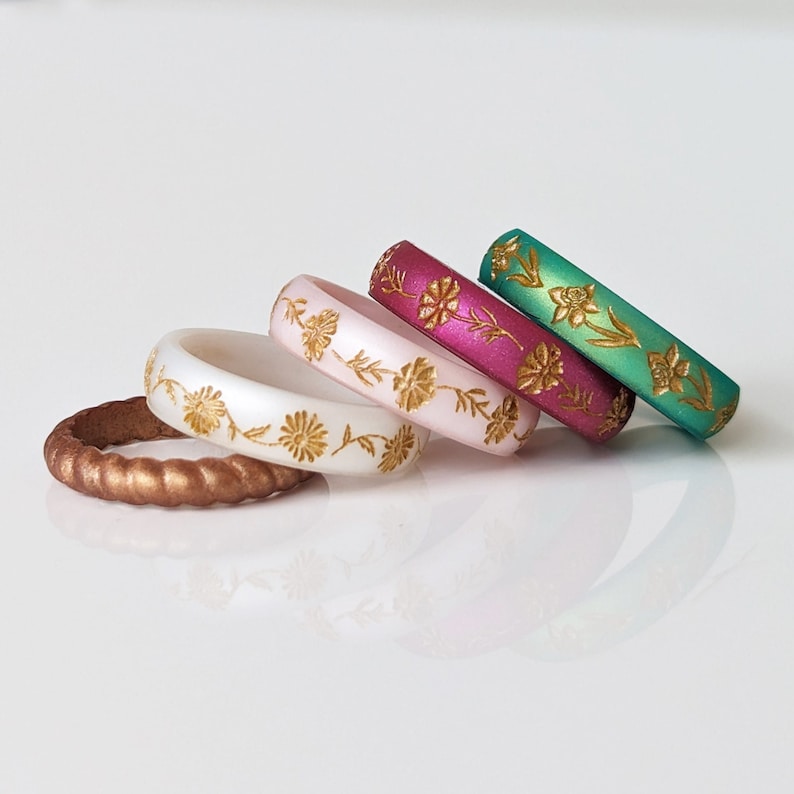 May include: Four silicone rings with a floral design. The rings are in shades of white, pink, red, and green. The floral design is in gold.