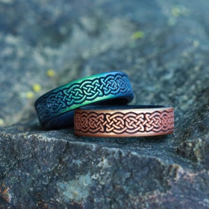 Op de afbeelding: Twee ringen met Keltische knoopmotieven. Eén ring heeft een blauw-groene gradiënt, de andere is bronskleurig. De ringen liggen op een grijze steen. De ringen zijn waarschijnlijk van metaal.