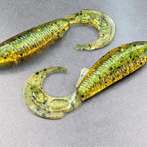 May include: Two soft plastic fishing lures. The lures are a mix of green and yellow with black speckles. They have a curved tail design. The lures are designed to attract fish.