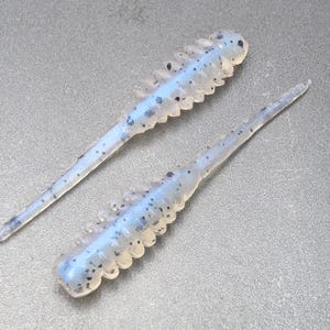 May include: Two soft plastic fishing lures with a translucent light blue body and small black speckles. Each lure features a segmented, ribbed upper section and a long, thin, tapered tail extending from the main body, designed for fishing.