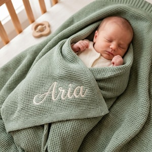 Könnte beinhalten: Ein schlafendes Neugeborenes, eingehüllt in eine salbeigrüne Waffelstrickdecke. Der Name "Aria" ist in weißer Schrift auf die Decke gestickt. Im Hintergrund ist ein Holzspielzeug zu sehen, was auf eine Kinderzimmereinrichtung hindeutet.