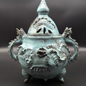 May include: A light blue ceramic incense burner with a lid, featuring intricate relief carvings of dragons on the body and handles. The vessel stands on three small feet and has a pointed lid with cut-out openings and dark brown accents.