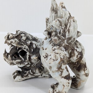 May include: A ceramic sculpture of a mythical creature, possibly a dragon or lion. The figure is primarily white with brown and black accents, featuring an open mouth with visible teeth and a textured, detailed body. The sculpture has a rough, aged appearance.