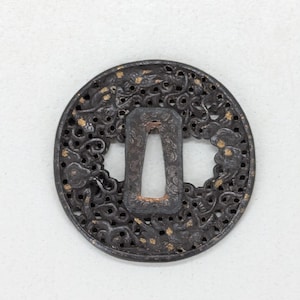 Op de afbeelding: Een ronde, donkere metalen tsuba, een Japanse zwaardbeschermer, met ingewikkelde opengewerkte ontwerpen. De beschermer heeft een centrale rechthoekige opening en is versierd met gouden accenten, met een gedetailleerd, sierlijk patroon.