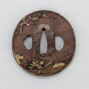 Op de afbeelding: Een ronde, koperkleurige metalen tsuba, een Japanse zwaardbeschermer, met ingewikkelde details. Het toont een krijgersfiguur en bloemmotieven in goud. De tsuba heeft een centrale opening en twee zijopeningen.