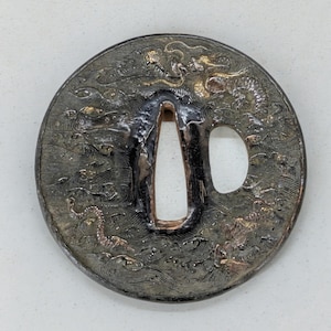 Op de afbeelding: Een cirkelvormige, antieke metalen tsuba met ingewikkelde reliëfsnijwerk. Het ontwerp toont een draak te midden van wervelende golven, met twee openingen voor een zwaard. De tsuba heeft een donkere, verouderde uitstraling met goudkleurige accenten.