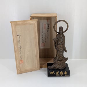 May include: A bronze-coloured statue of a robed figure with a halo, displayed on a black base with gold lettering. The statue is with a wooden box with Japanese text and a lid.