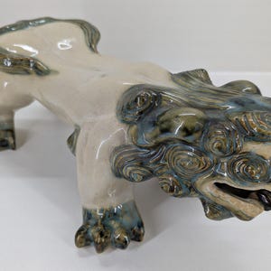 May include: A ceramic sculpture of a mythical creature, possibly a guardian lion. The figure is primarily off-white with accents of blue and green glaze. The creature is depicted in a crouching position with detailed features.