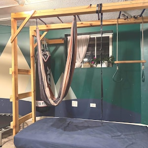 May include: A wooden climbing frame with exercise equipment, including a hanging hammock, rings, and a pull-up bar. The frame is positioned over a blue mattress. The background features a window and a painted wall with geometric shapes.