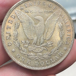Op de afbeelding: Een zilveren One Dollar munt met een adelaar met uitgespreide vleugels, omringd door tekst waaronder "UNITED STATES OF AMERICA" en "ONE DOLLAR". De munt heeft een gedetailleerd ontwerp en een licht versleten uiterlijk.