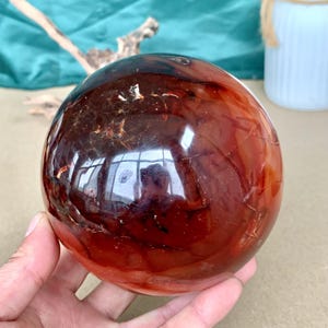 May include: A polished, spherical gemstone in shades of deep red and brown. The orb has a smooth, reflective surface with natural patterns and inclusions. The gemstone is held in a hand, showcasing its size and color variations.