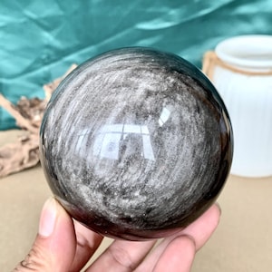 Könnte beinhalten: Eine polierte Obsidiankugel mit silbernem Glanz. Die Kugel hat ein wirbelndes Muster aus Silber und Schwarz. Die Kugel wird in einer Hand gehalten. Der Hintergrund umfasst einen türkisfarbenen Stoff, eine weiße Vase und ein Stück Treibholz.
