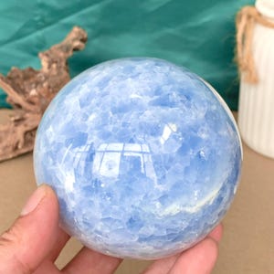 Puede incluir: Una gran esfera pulida de calcita azul. La bola de cristal tiene una apariencia nublada y texturizada con variaciones en el color azul. La esfera se sostiene en una mano, con un fondo borroso que incluye un jarrón blanco y madera flotante.