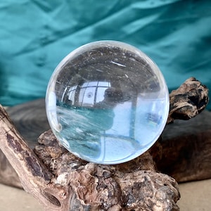 Könnte beinhalten: Eine klare, kugelförmige Kristallkugel liegt auf einem Stück verwittertem Holz. Die transparente Kugel reflektiert die Umgebung und erzeugt ein verzerrtes Bild. Das Holz hat eine raue, strukturierte Oberfläche mit verschiedenen Brauntönen.
