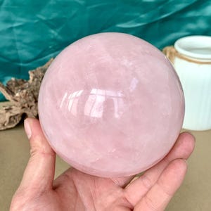 May include: A large, polished rose quartz sphere. The sphere is a soft pink color and reflects light, creating a glossy appearance. The crystal ball is held in a hand, with a blurred background.
