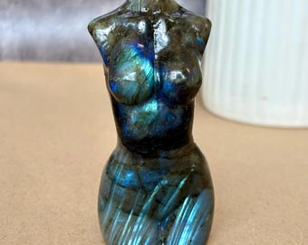 May include: A polished labradorite stone carving shaped like a human torso. The mineral displays a dark gray base color with iridescent flashes of electric blue and teal light reflecting across the chest, abdomen, and hip areas of the sculpture.