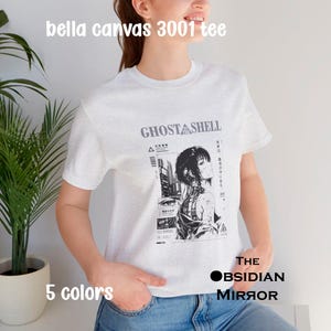 Könnte beinhalten: Ein hellgraues Kurzarm-T-Shirt mit einem schwarz-weißen Grafikdruck der Anime-Figur Ghost in the Shell. Das Design enthält japanischen Text, ein Stadtbild und den Titel Ghost in the Shell. Der Text bella canvas 3001 tee ist sichtbar.