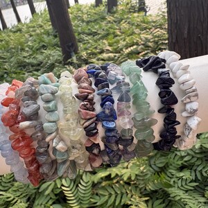 May include: A collection of gemstone chip bracelets in various colors, including red, blue, green, and white. The bracelets are strung together and displayed on a white surface, with a natural green background.