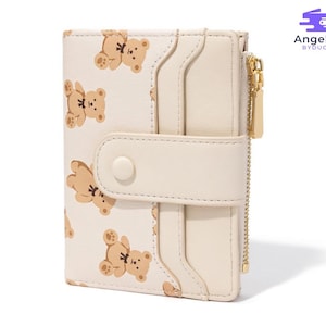 May include: Cream-colored bi-fold wallet with a repeating pattern of brown teddy bears. The wallet has a snap closure, card slots, and a gold zipper. The brand name "Angela BYDUC" is printed on the upper right corner.