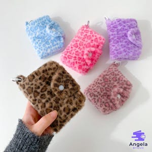 May include: A collection of small, fluffy wallets in various colors, including blue, pink, purple, brown, and mauve. Each wallet features a leopard print design and a button closure. The wallets are attached to silver keychains.