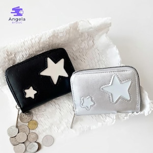 May include: Two small, zippered wallets with star designs. One wallet is black with white stars, and the other is silver with white stars. The wallets are displayed on a white, textured surface, with coins scattered nearby. The brand name "Angela BYDUC" is visible.