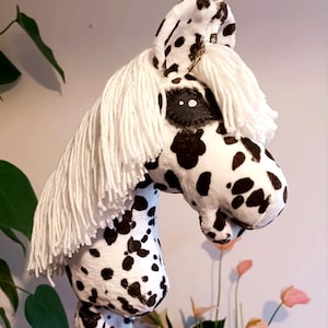 Peut inclure: Un cheval bâton en peluche avec un corps en tissu blanc à taches noires, une longue crinière en laine blanche et des oreilles droites. La tête du cheval est faite d'un matériau souple et présente une tache sombre autour de l'œil.