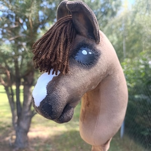 Peut inclure: Un cheval bâton en peluche avec une tête en tissu marron, une crinière en laine marron foncé, une tache blanche sur le museau et des yeux cousus noirs. La tête de cheval est fixée sur un bâton en bois, avec des coutures détaillées.