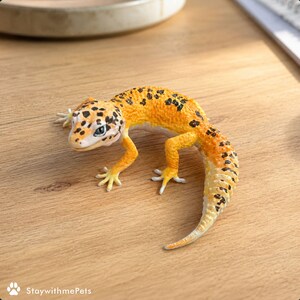 Pode incluir: Figura detalhada de um gecko leopardo, com um corpo laranja e amarelo e manchas pretas. O gecko tem uma cabeça com padrões brancos e pretos, colocada sobre uma superfície de madeira. O item é temático de animais de estimação.