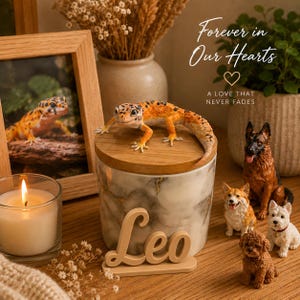 Könnte beinhalten: Ein marmoriertes Keramikgefäß mit Holzdeckel, gekrönt von einer Leopardgecko-Figur. Das Gefäß trägt den Namen Leo in Schreibschrift. Daneben stehen Figuren eines Deutschen Schäferhunds, eines Corgis, eines Westies und eines Pudels.