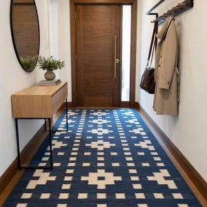 Op de afbeelding: Een rechthoekig vloerkleed met een diepblauwe achtergrond en een herhalend wit geometrisch patroon van kruisen en vierkanten. Het kleed ligt in een hal met een houten deur, een sidetable van licht hout, een ronde spiegel en een wandkapstok.