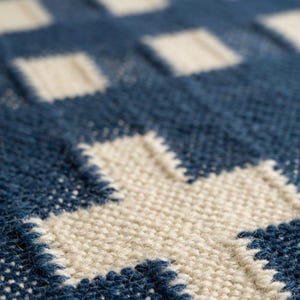 Op de afbeelding: Een close-up van een geweven textiel met een herhalend geometrisch patroon. De stof toont een raster van donkerblauwe en gebroken witte vierkanten, met een opvallend kruisvormig motief gevormd door de lichte draden tegen de donkerblauwe achtergrond.