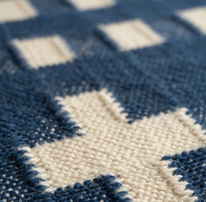 Op de afbeelding: Een close-up van een geweven textiel met een herhalend geometrisch patroon. De stof toont een raster van donkerblauwe en gebroken witte vierkanten, met een opvallend kruisvormig motief gevormd door de lichte draden tegen de donkerblauwe achtergrond.