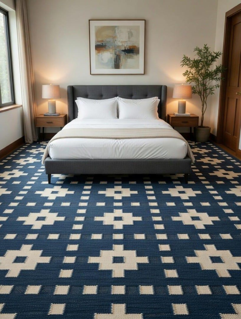 Op de afbeelding: Een groot vloerkleed met een diepblauwe achtergrond en een herhalend geometrisch kruispatroon in cr&egrave;mekleur. Het kleed bedekt de vloer van een slaapkamer en strekt zich uit van de voorgrond naar een grijs gestoffeerd bedframe met wit beddengoed.