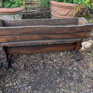 May include: A rustic wooden planter, featuring a rectangular trough design, set on a dark-stained wooden stand. The planter is filled with soil and greenery. The wood shows visible grain and knots, suggesting an aged, outdoor aesthetic.