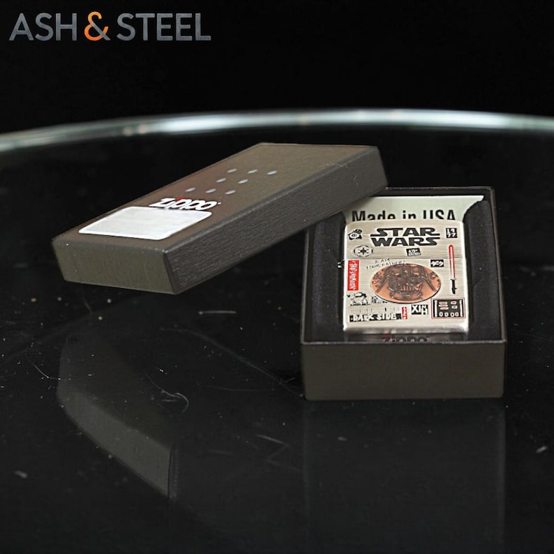 K&ouml;nnte beinhalten: Ein silbernes Zippo-Feuerzeug mit Star Wars-Design, pr&auml;sentiert in einer schwarzen Box. Das Feuerzeug zeigt verschiedene Star Wars-Grafiken und den Text "Made in USA". Die Box ist ge&ouml;ffnet und zeigt das Feuerzeug.