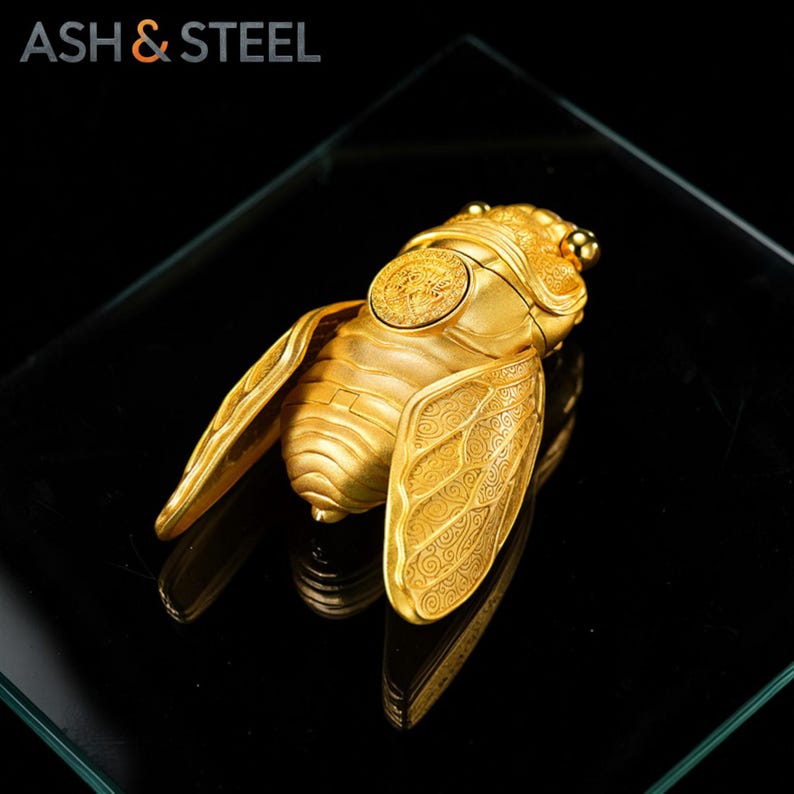 Puede incluir: Un objeto dorado con forma de cigarra, con intrincados detalles en las alas y un emblema circular en la parte posterior. Se muestra sobre una superficie negra reflectante. El texto "ASH & STEEL" es visible.